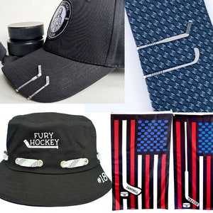 May include: A black baseball cap with a white logo that says "Fury Hockey" and a hockey stick graphic. The cap has a white band with a pattern and a white number "18" on the side. There are two silver hockey stick tie clips on the cap. There are also two American flag designs with hockey sticks and the names "Campbell" and "Traver" on them.