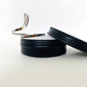 May include: A silver, open-ended bangle bracelet with a textured design. The bracelet is displayed in a black round tin with a lid.