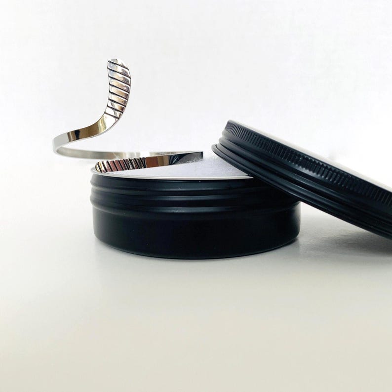 May include: A silver, open-ended bangle bracelet with a textured design. The bracelet is displayed in a black round tin with a lid.