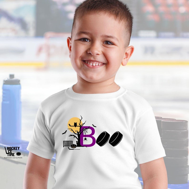 May include: A white t-shirt with a purple letter "B" and two black hockey pucks. The letter "B" is surrounded by a Halloween-themed design with a tree, bats, an owl, and a spider web.