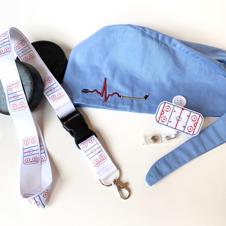 May include: A white lanyard with a hockey rink design, a black buckle, and a silver clasp. A blue scrub cap with a hockey-themed heartbeat design and a hockey rink badge are also present, along with a hockey puck.