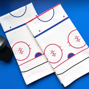 May include: Two white cotton tea towels with a hockey rink design. The rink is outlined in black and red, with blue circles for the face-off dots. The towels are folded in half and placed on a blue surface. Two black hockey pucks are in the lower left corner of the image.