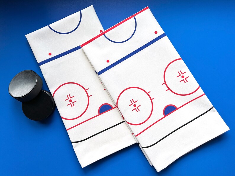 May include: Two white cotton tea towels with a hockey rink design. The rink is outlined in black and red, with blue circles for the face-off dots. The towels are folded in half and placed on a blue surface. Two black hockey pucks are in the lower left corner of the image.