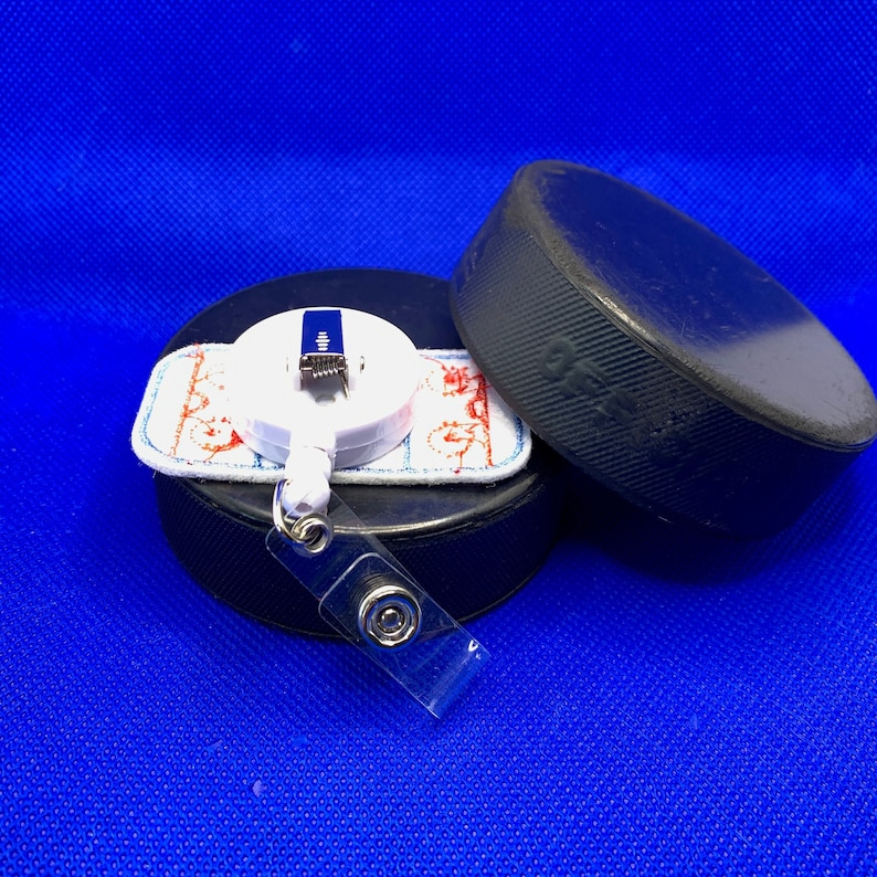 May include: A white retractable badge reel with a hockey-themed design, featuring a hockey puck and a clear plastic strap. The reel is placed on top of a black hockey puck, with another puck in the background. The background is a solid blue.