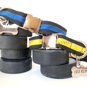 May include: Two dog collars made from hockey laces. One collar is black with blue stripes and the other is black with yellow stripes. Both collars have silver buckles and bone-shaped tags that read "Lost as Puck". The collars are displayed on top of hockey pucks.
