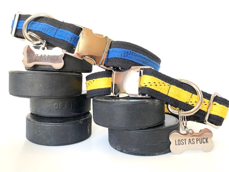 May include: Two dog collars made from hockey laces. One collar is black with blue stripes and the other is black with yellow stripes. Both collars have silver buckles and bone-shaped tags that read "Lost as Puck". The collars are displayed on top of hockey pucks.