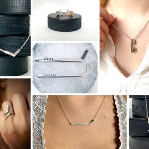 May include: A set of hockey-themed jewelry, including a necklace with a hockey stick pendant, a ring with a hockey stick design, and a pair of earrings with a hockey puck design. The jewelry is displayed on a black hockey puck.