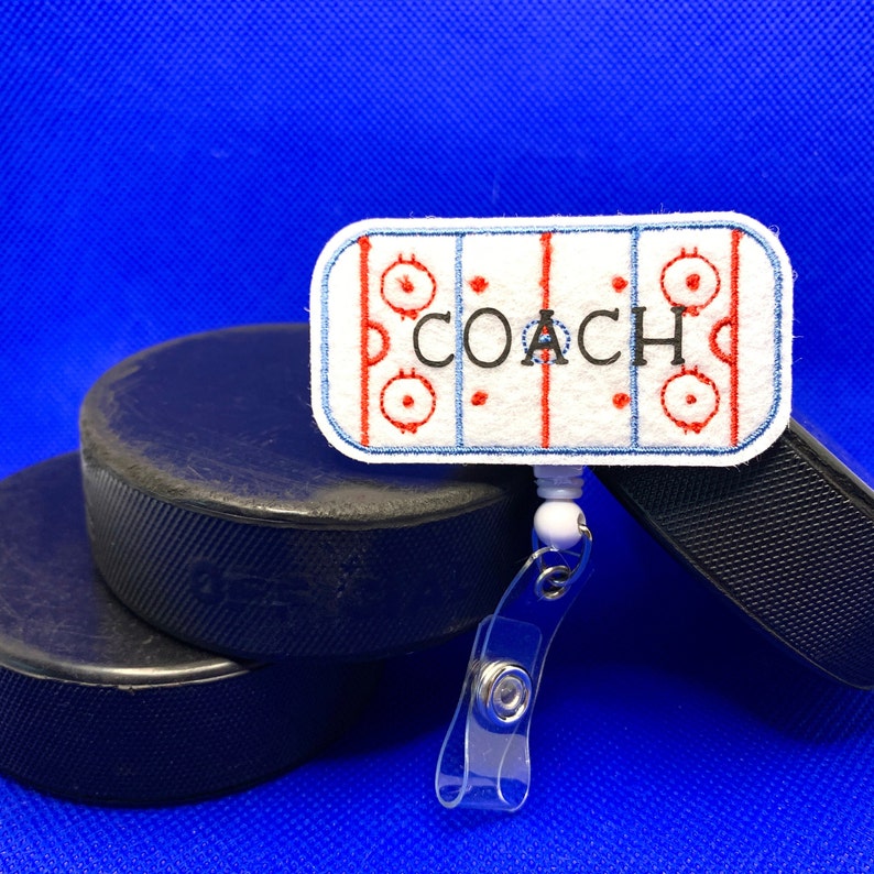 May include: A white and red hockey rink badge reel with the word "COACH" in black letters. The badge reel is attached to a clear plastic clip. Two black hockey pucks are in the background.