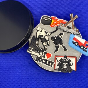 May include: A silver charm with a hockey theme. The charm features a hockey player, a hockey stick, a puck, a net, and the words "I love hockey".