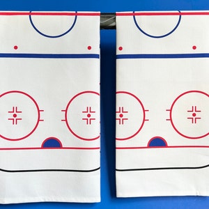 May include: Two white fabric kitchen towels with a hockey rink design printed on them. The rink is outlined in red and blue, with a center circle and two face-off circles. The towels are hanging from a metal rod.