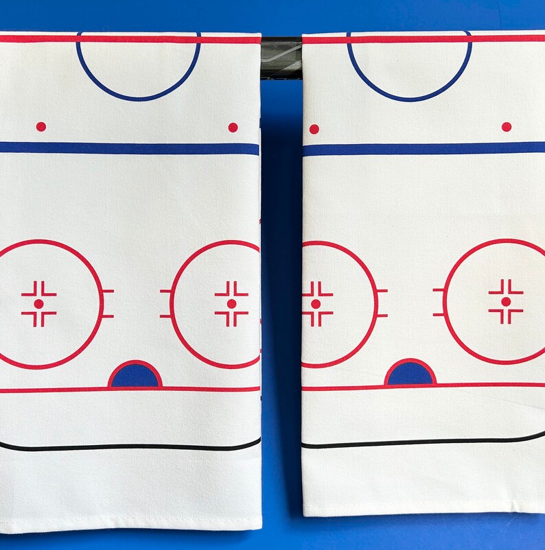 May include: Two white fabric kitchen towels with a hockey rink design printed on them. The rink is outlined in red and blue, with a center circle and two face-off circles. The towels are hanging from a metal rod.