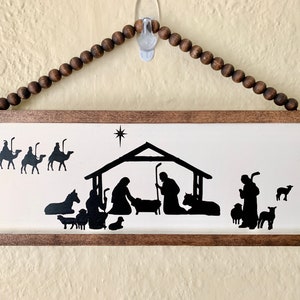 May include: A wooden framed sign with a white background and a black silhouette of the nativity scene. The three wise men are on camels in the top left corner, and a shepherd with sheep is on the right. The star of Bethlehem is above the manger.
