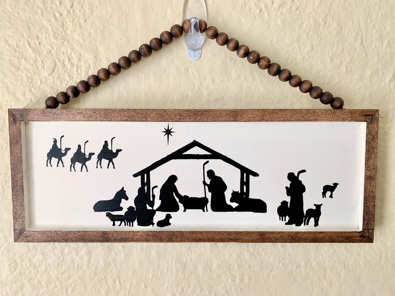 May include: A wooden framed sign with a white background and a black silhouette of the nativity scene. The three wise men are on camels in the top left corner, and a shepherd with sheep is on the right. The star of Bethlehem is above the manger.