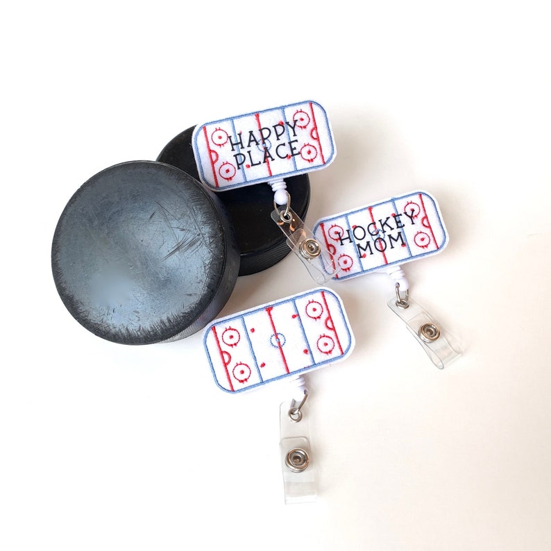 May include: Three hockey-themed retractable badge holders with a hockey rink design and the phrases "Happy Place" and "Hockey Mom". Two black hockey pucks are also in the image. The badge holders have a white background with red and blue details.