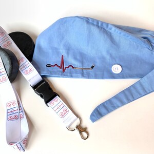 May include: A light blue scrub cap with a hockey-themed design, featuring an embroidered heartbeat and hockey stick. A white lanyard with a hockey rink pattern and a black buckle is also present, along with a hockey puck.