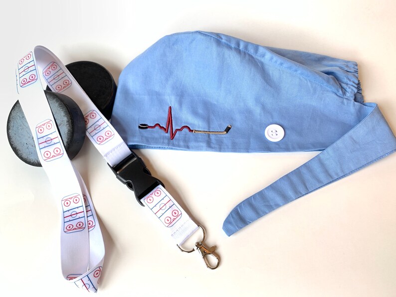 May include: A light blue scrub cap with a hockey-themed design, featuring an embroidered heartbeat and hockey stick. A white lanyard with a hockey rink pattern and a black buckle is also present, along with a hockey puck.
