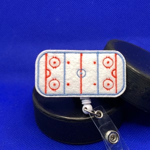 May include: A white felt hockey rink badge reel with red and blue embroidered details. The badge reel is attached to a clear plastic holder and is resting on top of a black hockey puck. The background is a solid blue.