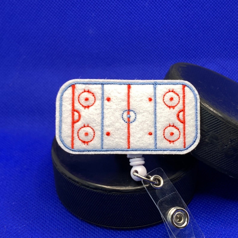 May include: A white felt hockey rink badge reel with red and blue embroidered details. The badge reel is attached to a clear plastic holder and is resting on top of a black hockey puck. The background is a solid blue.