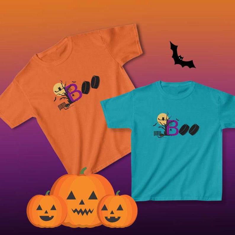 May include: Two t-shirts with a Halloween theme. The orange shirt has a purple "B" and the blue shirt has a purple "B" with a black "OO" behind it. Both shirts have a black silhouette of a tree with bats and a moon. The shirts are displayed with three carved pumpkins.