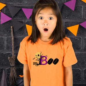 May include: A young person wearing an orange t-shirt with a Halloween-themed design featuring a purple letter "B", a black tree, a black owl, a black spider, a black hockey net, a black hockey stick, and two black hockey pucks. The design says "BOO".