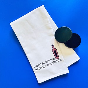 May include: White kitchen towel with the text "I can't talk right now, I'm doing Hockey Mom shit." and a pink glitter wine bottle graphic.