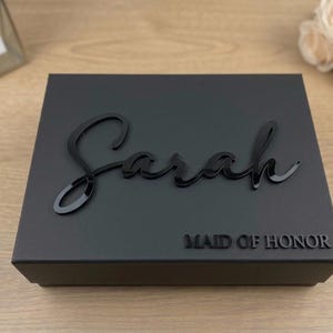 May include: A rectangular matte black gift box featuring the name Sarah in a glossy black script font on the lid. Below the name, the text MAID OF HONOR is displayed in a clean, sans-serif black font. The box has a structured, flat design.