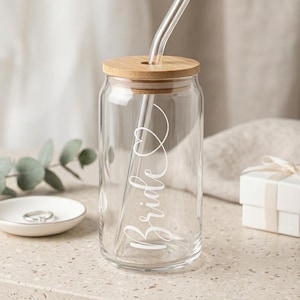 Könnte beinhalten: Ein durchsichtiges Glas in zylindrischer Form mit dem weißen, kursiven Schriftzug "Bride" und einem Herz-Umriss. Das Glas verfügt über einen hellen Bambusdeckel mit einer runden Öffnung und einen wiederverwendbaren, durchsichtigen Glasstrohhalm.