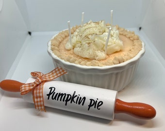 May include: A white ceramic pie dish with a pumpkin pie candle inside. The candle has a brown crust, orange filling, and white whipped cream topping. A white rolling pin with the words "Pumpkin Pie" written on it is in the foreground.