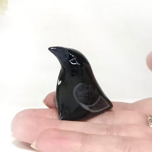 May include: A small, glossy black bird ornament with a white heart detail on its chest, resting on a person's open palm. A vase of yellow and white flowers is in the background.