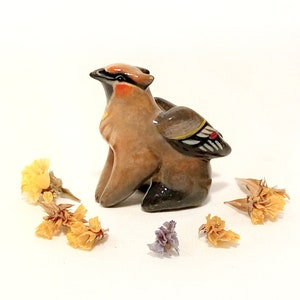 May include: A ceramic figurine of a brown and black bird with a red and white wing. The bird is sitting on a white surface with dried flowers around it.