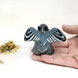 May include: A small, grey ceramic angel figurine with blue wings and black and white striped details. The angel is held in a hand, with dried flowers in the background. The figurine has a glossy finish.
