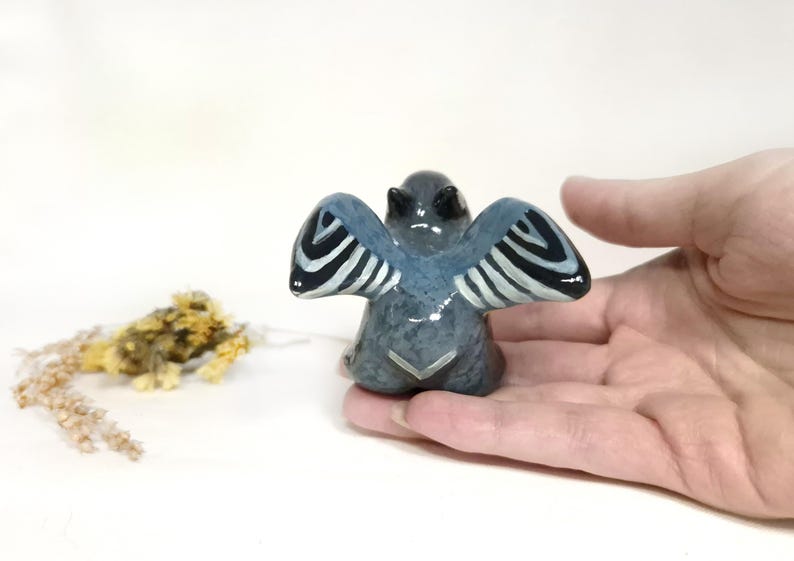 May include: A small, grey ceramic angel figurine with blue wings and black and white striped details. The angel is held in a hand, with dried flowers in the background. The figurine has a glossy finish.