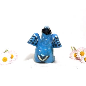 May include: A small, blue ceramic bird figurine with black and white wing details. The bird is positioned in front of a white background, with small white and yellow flowers on either side.