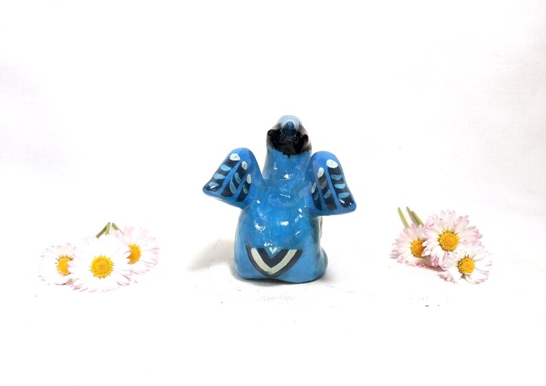 May include: A small, blue ceramic bird figurine with black and white wing details. The bird is positioned in front of a white background, with small white and yellow flowers on either side.