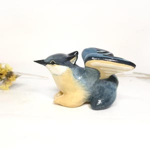 May include: A ceramic bird figurine in shades of blue, beige, and black, with outstretched wings. The bird is perched, with its wings spread, and has a detailed beak. Dried yellow flowers are in the background.
