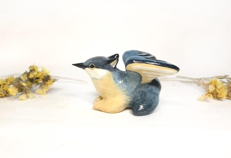May include: A ceramic bird figurine in shades of blue, beige, and black, with outstretched wings. The bird is perched, with its wings spread, and has a detailed beak. Dried yellow flowers are in the background.