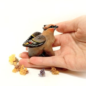 May include: A small ceramic figurine of a brown and black bird with yellow and red accents on its wings. The bird is perched on a hand.
