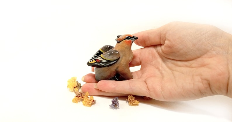 May include: A small ceramic figurine of a brown and black bird with yellow and red accents on its wings. The bird is perched on a hand.