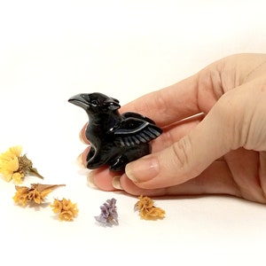 May include: Black obsidian crow figurine with wings outstretched. The figurine is about 2 inches tall and is sitting on a white surface.