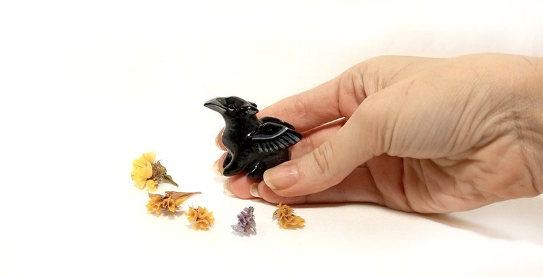 May include: Black obsidian crow figurine with wings outstretched. The figurine is about 2 inches tall and is sitting on a white surface.
