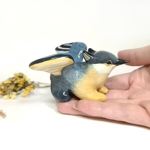 May include: A hand-painted ceramic bird figurine with outstretched wings. The bird has a blue and grey body with yellow underparts and wings. The beak is black. A small sprig of dried flowers is in the background.