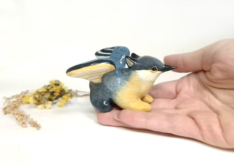 May include: A hand-painted ceramic bird figurine with outstretched wings. The bird has a blue and grey body with yellow underparts and wings. The beak is black. A small sprig of dried flowers is in the background.