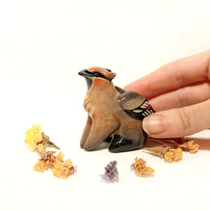 May include: A small ceramic figurine of a brown and black bird with a red head and a white and black wing. The figurine is sitting on a white surface with dried flowers around it.