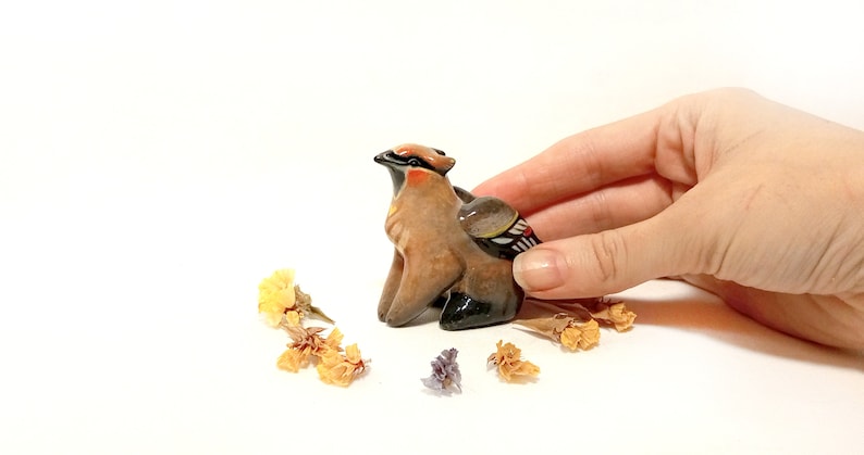 May include: A small ceramic figurine of a brown and black bird with a red head and a white and black wing. The figurine is sitting on a white surface with dried flowers around it.