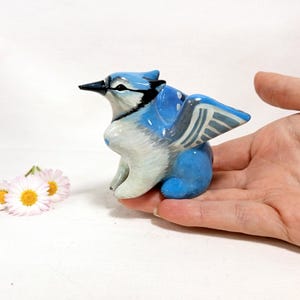 May include: A hand-painted ceramic blue jay figurine with white and black accents. The bird has detailed wings and a black beak. Two small daisies with yellow centers are next to the figurine.