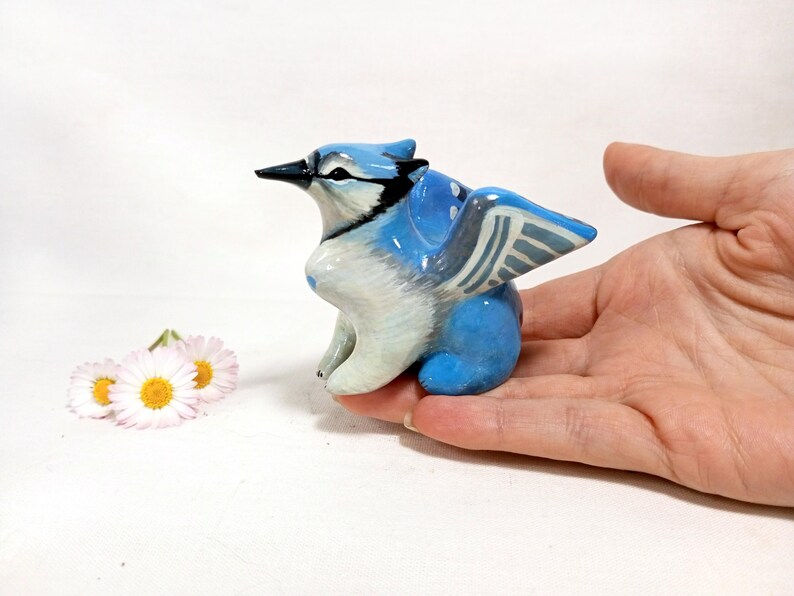 May include: A hand-painted ceramic blue jay figurine with white and black accents. The bird has detailed wings and a black beak. Two small daisies with yellow centers are next to the figurine.