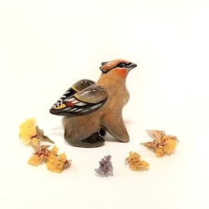 May include: A small ceramic figurine of a brown and black bird with yellow, red, and white markings on its wings. The bird is standing on a white surface with dried yellow flowers around it.