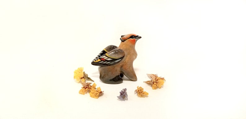 May include: A small ceramic figurine of a brown and black bird with yellow, red, and white markings on its wings. The bird is standing on a white surface with dried yellow flowers around it.