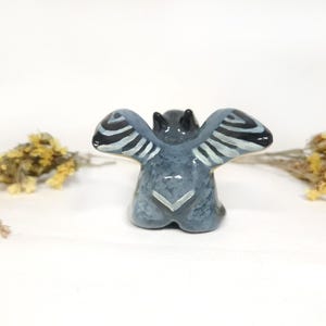 May include: A ceramic bat figurine, viewed from the back, with outstretched wings. The bat is painted in shades of grey and blue, with black and white striped wing details. The figurine is on a white surface.