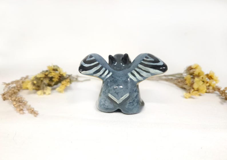 May include: A ceramic bat figurine, viewed from the back, with outstretched wings. The bat is painted in shades of grey and blue, with black and white striped wing details. The figurine is on a white surface.